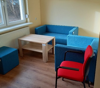 Pokój indywidualnych konsultacji w Centrum Wsparcia. Po środku stoi niski drewniany stolik, a wokół niego stoją niebieskie meble: kanapa, fotel i pufa oraz czerwone krzesło.