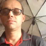 Pan Paweł Słowik w okularach, ubrany w szarą bluzę. Trzyma za sobą rozłożony parasol.