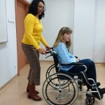 Na zdjęciu w sali wykładowej dwie kobiety, jedna siedzi na wózku inwalidzkim druga pcha wózek. Obie kobiety są uczestniczkami szkolenia, doświadczają niepełnosprawności.