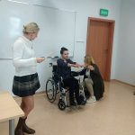 Na zdjęciu w sali wykładowej trzy kobiety, jedna siedzi na wózku inwalidzkim z wyciągniętymi rękoma do przodu, druga pochylona z miarką mierząca zasięg jej rąk, trzecia- prowadząca szkolenie- instruuje jak wykonać pomiar.