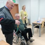 Na zdjęciu w sali wykładowej osoba prowadząca szkolenie kobieta oraz dwóch mężczyzn, jeden z nich na wózku inwalidzkim, w ramach szkolenia doświadcza niepełnosprawności.