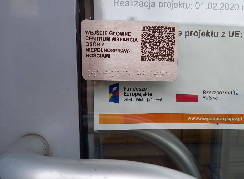 Fragment drzwi z klamką. Nad klamką tabliczka informacyjna z kodem qr oraz opisem w języku Braille’a. Za tabliczką fragment plakatu promującego projekt unijny.