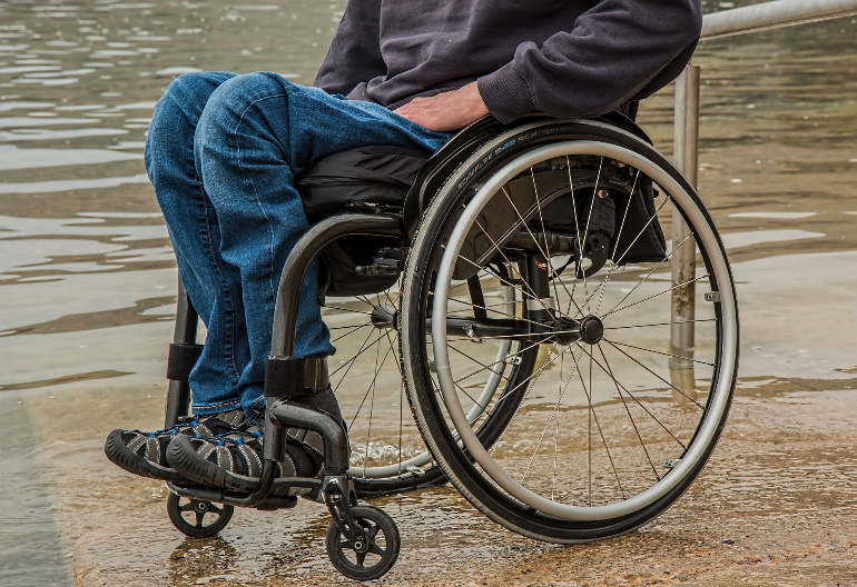Wózek inwalidzki, na którym siedzi mężczyzna ubrany w szarą kurtkę i dżinsowe spodnie. Wózek stoi nad wodą.