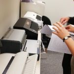 Na biurku stoją drukarki brailowskie i urządzenia do bindowania. Obok pracownik tłumaczy uczestnikowi wizyty działanie urządzenia.