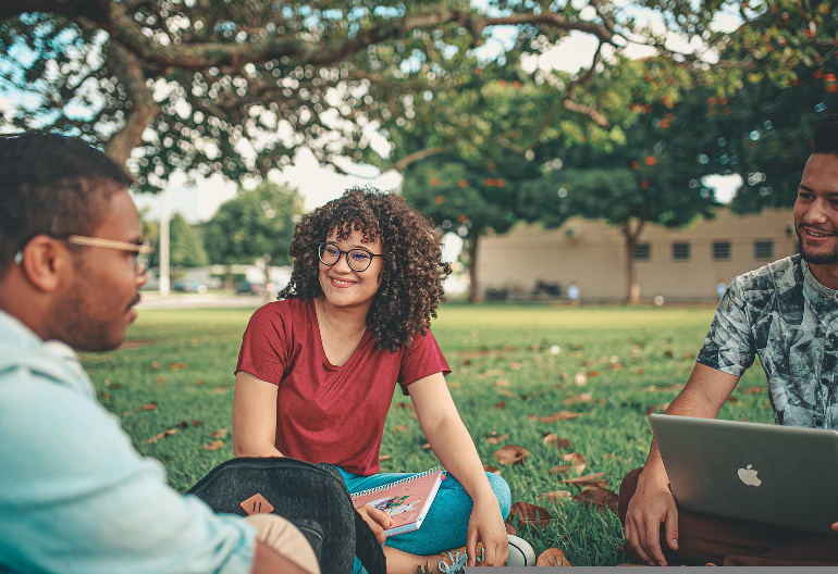 Troje studentów rozmawia siedząc na trawie przed budynkiem. Jeden z nich ma przed sobą laptopa.