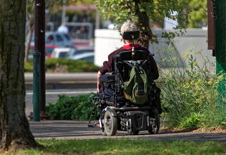 Elektryczny wózek inwalidzki, którym po chodniku w parku przemieszcza się osoba o siwych włosach w ciemnej bluzie. Z tyłu wózka jest plecak.