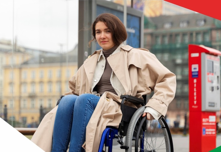 Kobieta ubrana w jasny płaszcz siedzi na wózku inwalidzkim trzymając obiema rękoma za koła. W tle jest zabudowa miasta.