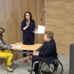 Pani doktor Justyna Palacz podsumowuje rozmowę siedzących przy stoliku prelegentów trzymając w ręku mikrofon i teczkę.