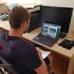 Stanowisko gry terenowej. Student, siedzi w pracowni przy biurku, przed sobą ma laptopa i za pomocą urządzenia PC Eye rozwiązuje zagadkę.